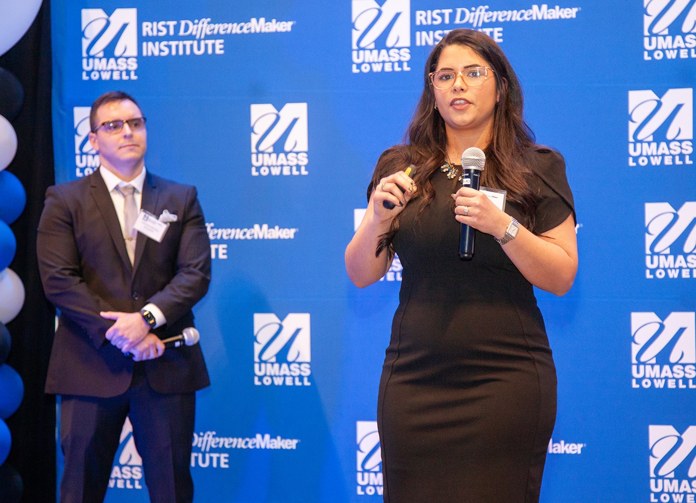 Ariel Pena-Martinez, computer engineering graduate student, holding a microphone and speaking in front of blue UMass Lowell backdrop while Grissel Cervantes-Jaramillo looks on.