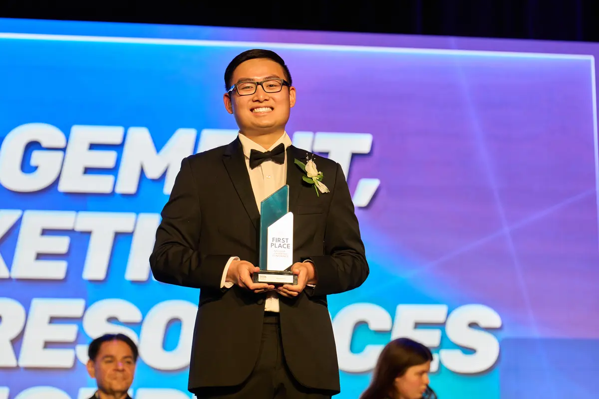 A young man in a suit with a bow tie smiles while holding a first place trophy on a stage.