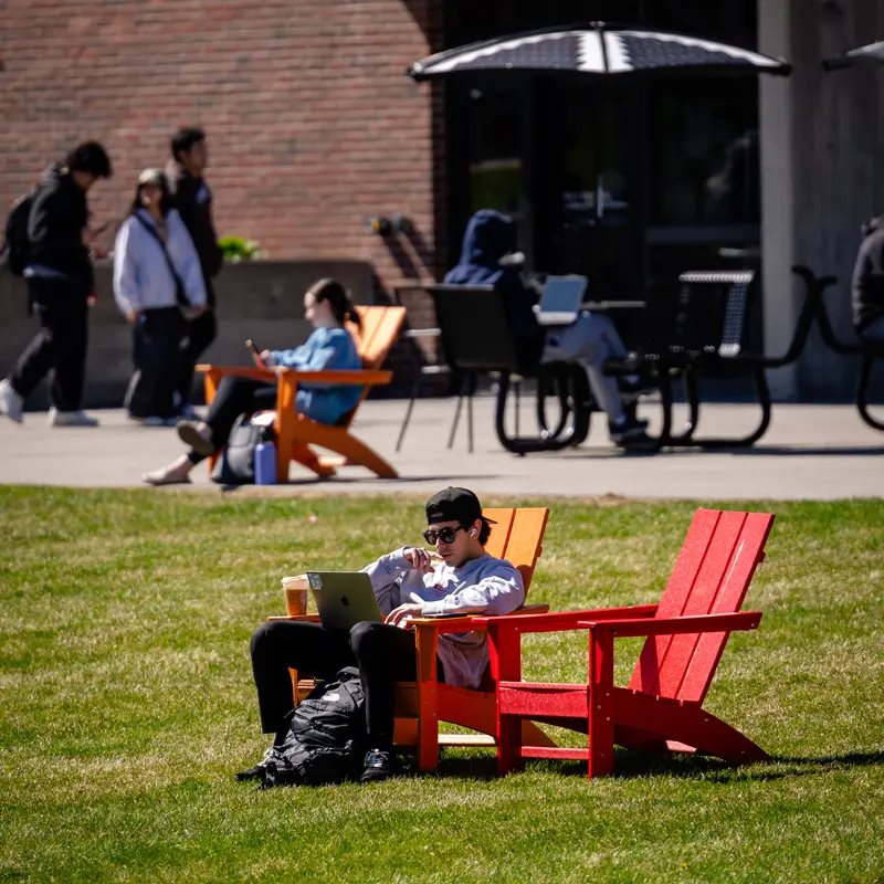 student sitting in red adirondack chair working on laptop on UMass Lowell's South Campus.
