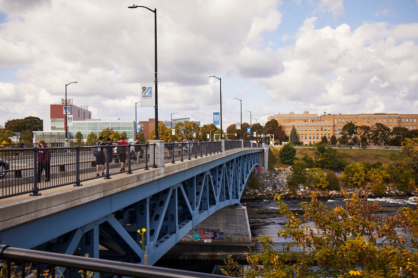 A bridge is shown from the side with a river flowing underneath. Several students are shown walking across the bridge. The bridge also features several white and blue UMass Lowell banners. Several UMass Lowell buildings are shown in the background