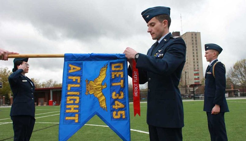 Tyler Davis is wearing air force uniform as he ties a red banner to a flag that displays a golden eagle and the words "DET 345 A FLIGHT."