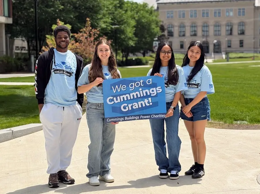 Four students stand on the south quad displaying a sign that reads We Got a Cummings Grant