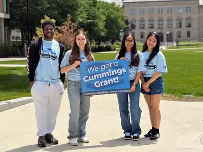 Four students stand on the south quad displaying a sign that reads We Got a Cummings Grant