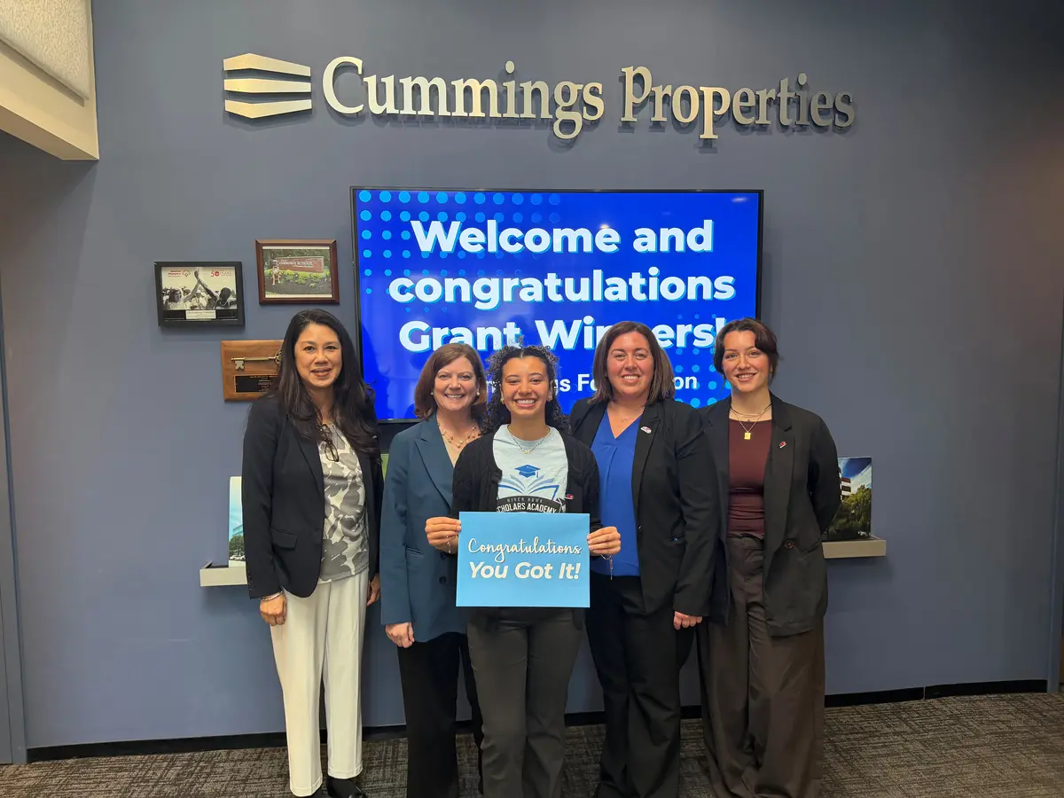 Five people in business attire in the foyer of Cummings Properties in front of a screen that says Welcome and Congratulations Grant Winners and holding a sign that says Congratulations You Got It