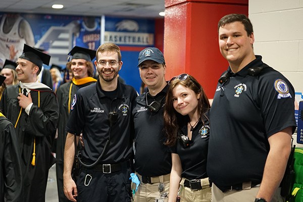 Four EMS workers pose for a photo while college graduates walk behind them