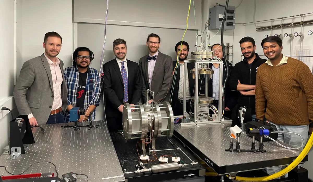 Eight researchers gather around a constant volume combustion chamber.
