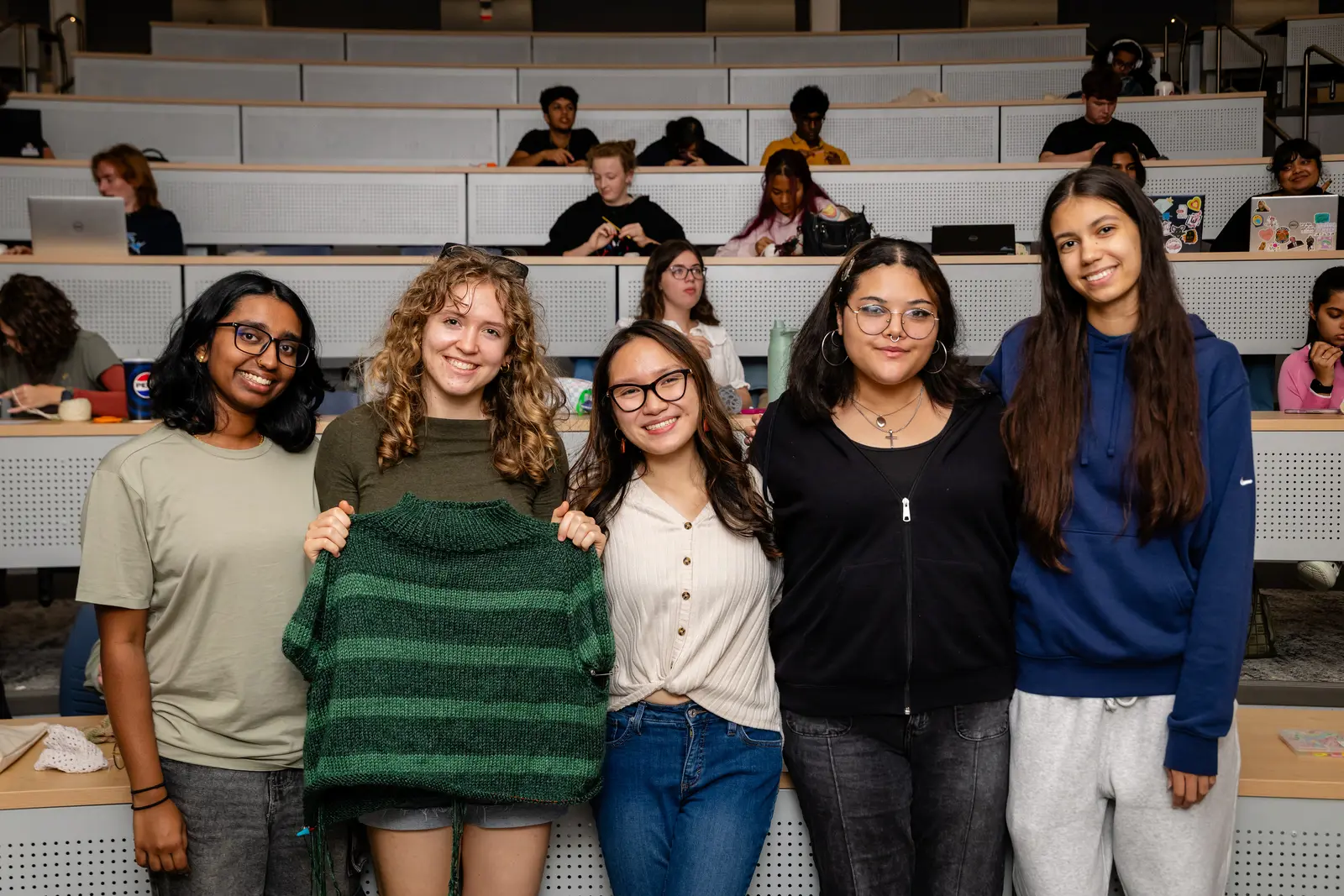Five students standing together smiling, one holding a crocheted sweater. 