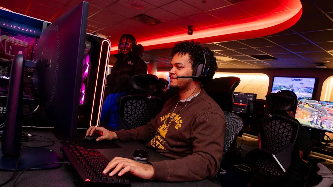 Student sits at gaming console wearing a headset while other student stand in background