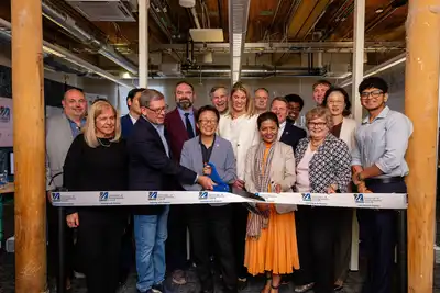Sixteen people line up for a photo behind a white ribbon stretched beneath two wooden pillars. The person in the middle holds an oversized pair of scissors