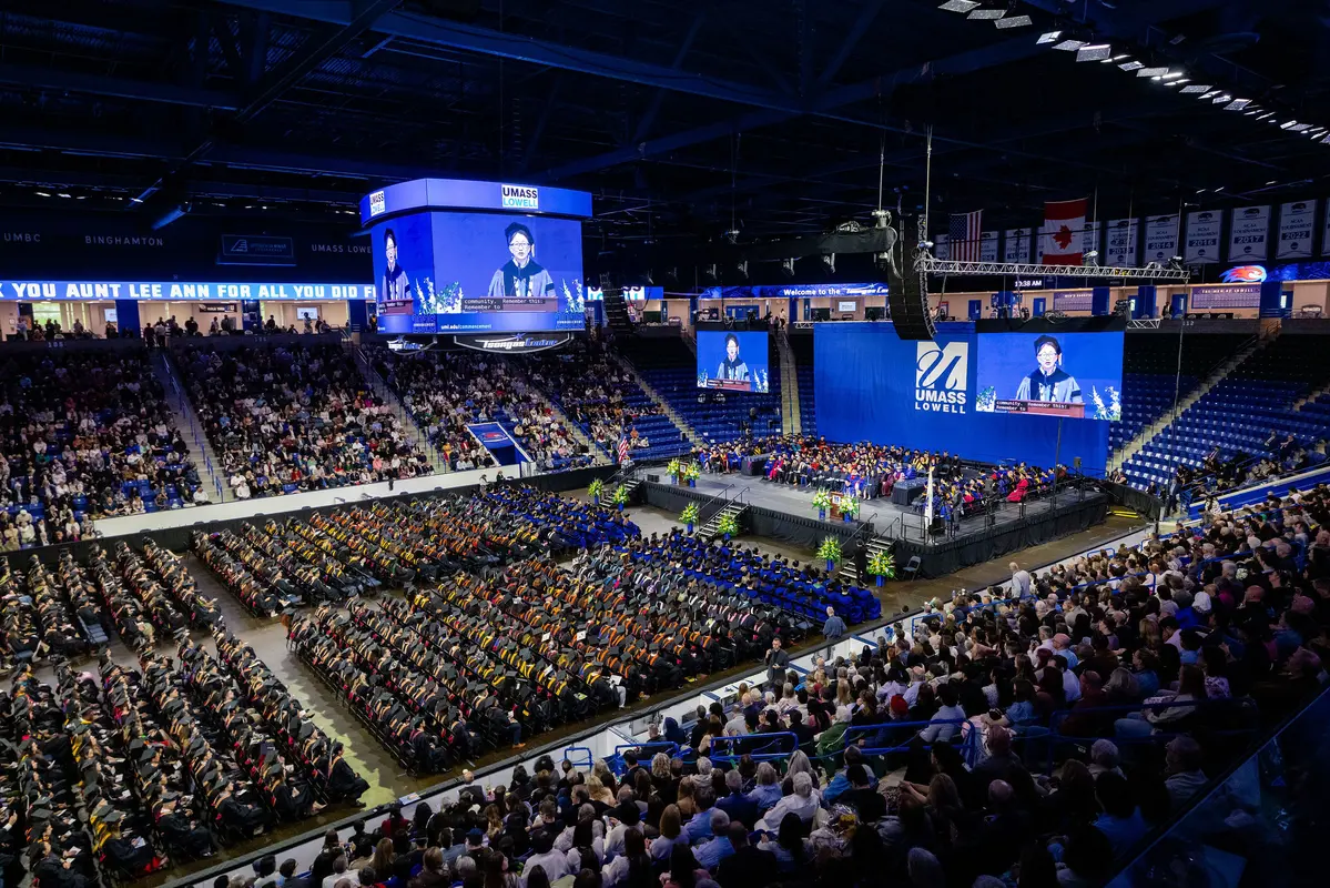 A wide shot of the floor of the Tsongas Center shows thousands of UMass Lowell graduates in their commencement regalia with families in the chairs surrounding them