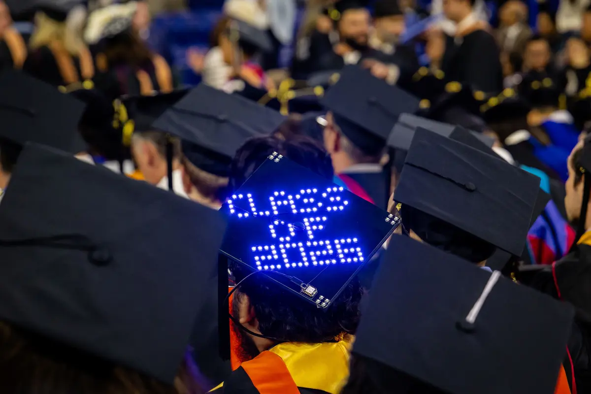 A single black mortar board in a sea of mortar boards is decorated with the illuminated words Class of 2026 