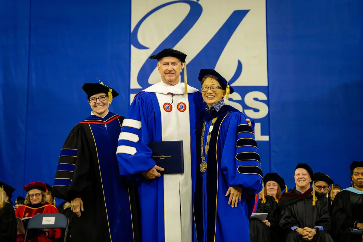 Vice Chancellor Anne Maglia, retired Brigadier General Jack Hammond and Chancellor Julie Chen pose for a photo in their commencement regalia in front of a blue and white UMass Lowell logo
