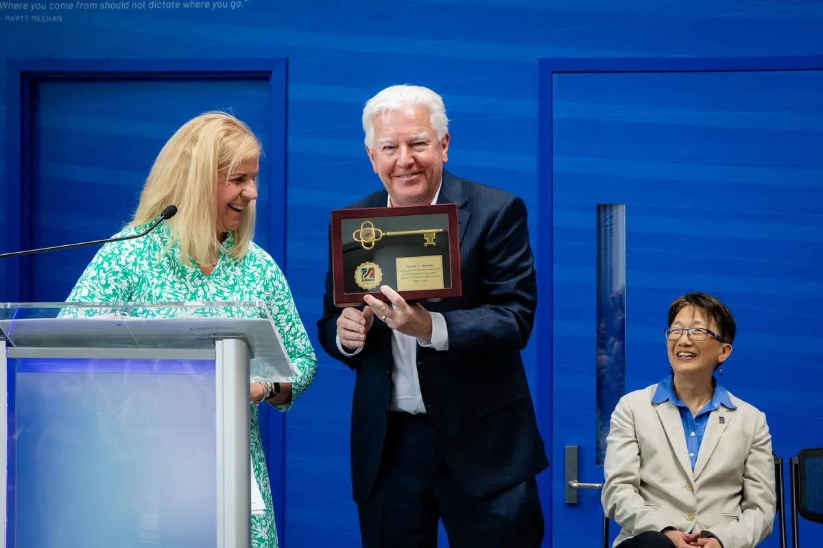 President Marty Meehan holds a plaque showcasing a golden key next to Trustee Mary Burns standing at a podium while Chancellor Chen looks on from her seat