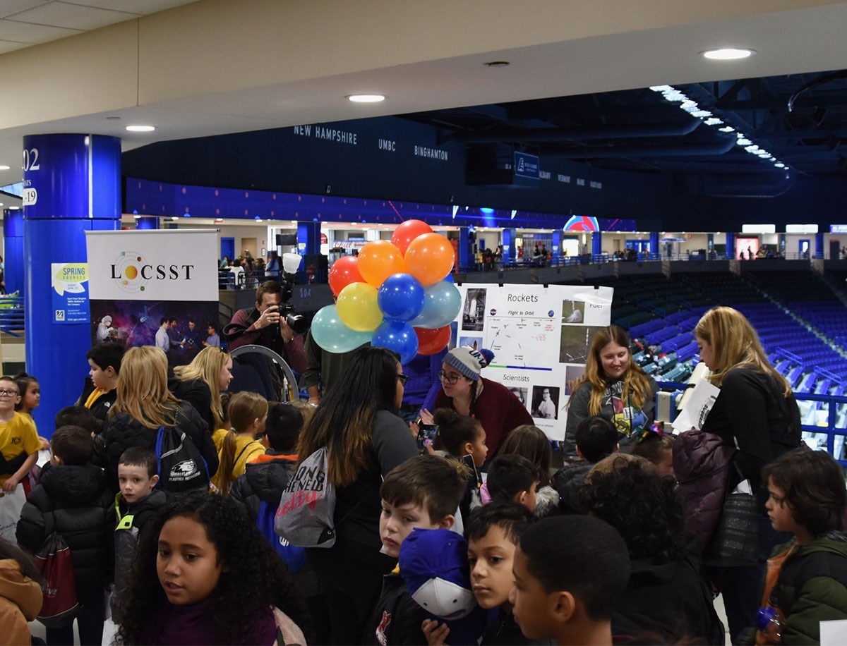 Young students are crowded around the LoCCST table at the UMass Lowell Field Trip Day. There are balloons on the table. 