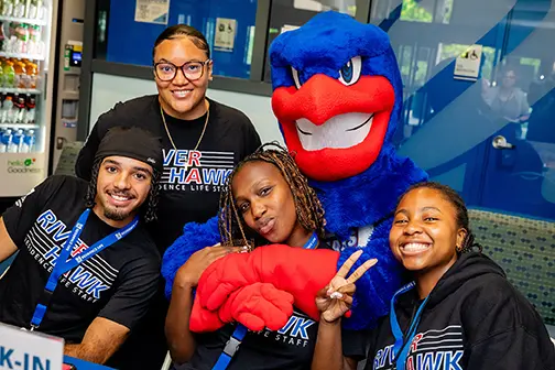 Photo shows UMass Lowell students at table in Fox Hall surrounding university mascot, Rowdy the River Hawk