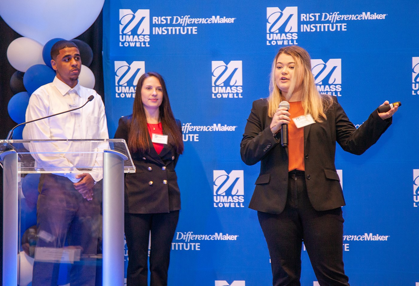 One of the members USuccess team holding a microphone and speaking in front of blue UMass Lowell backdrop while two other members watch.