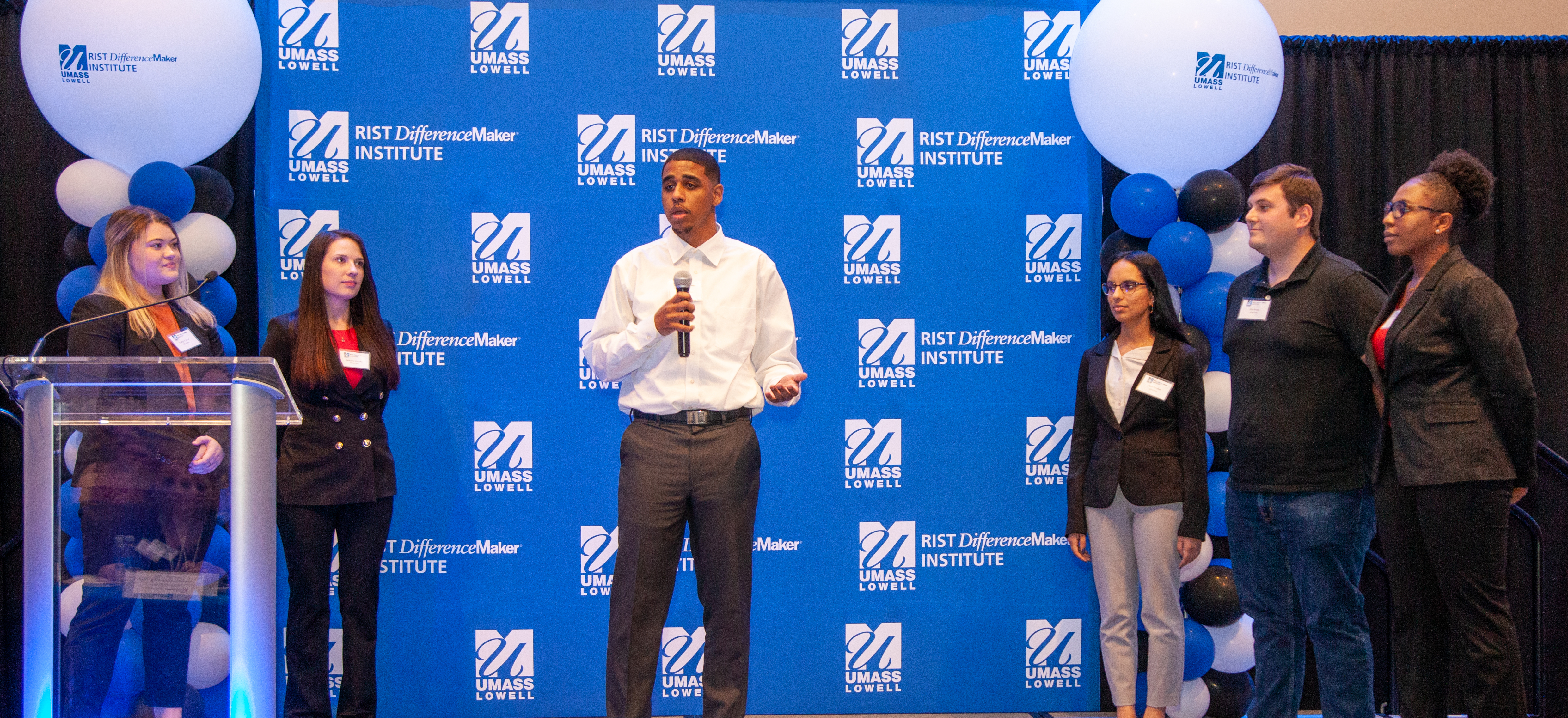 One of the members USuccess team holding a microphone and speaking in front of blue UMass Lowell backdrop while 5 others look on.