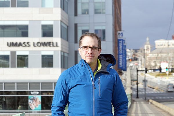 A man in glasses and a blue jacket stands on a bridge with a building behind him