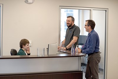 Two men stand at a reception desk and talk to a person who is seated