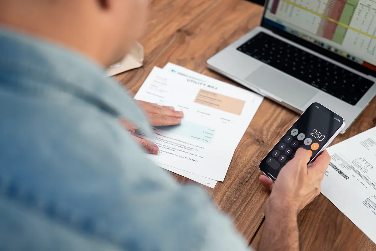 A man holds his utility bill, adding $250 to his calculator in front of a budget excel sheet.