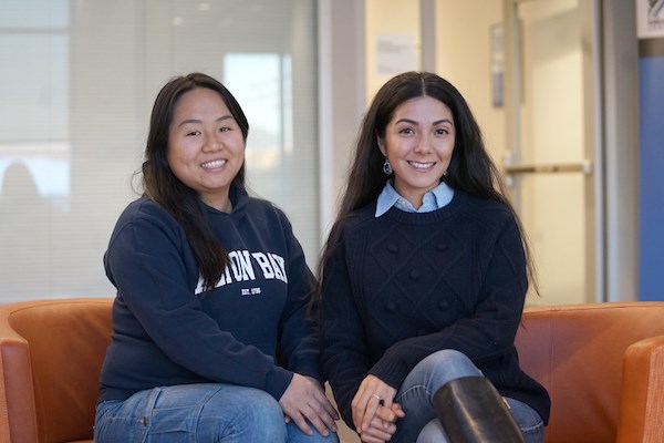 Two students with long dark hair smile while sitting in orange chairs and posing for a photo