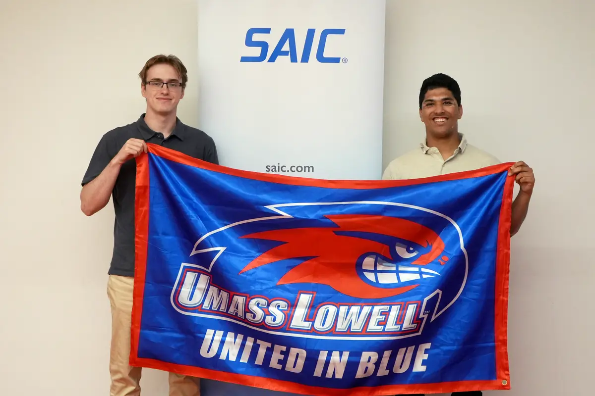 Viktor Akhonen and Rohan Paranjape hold a UMass Lowell flag in front of an SAIC sign.