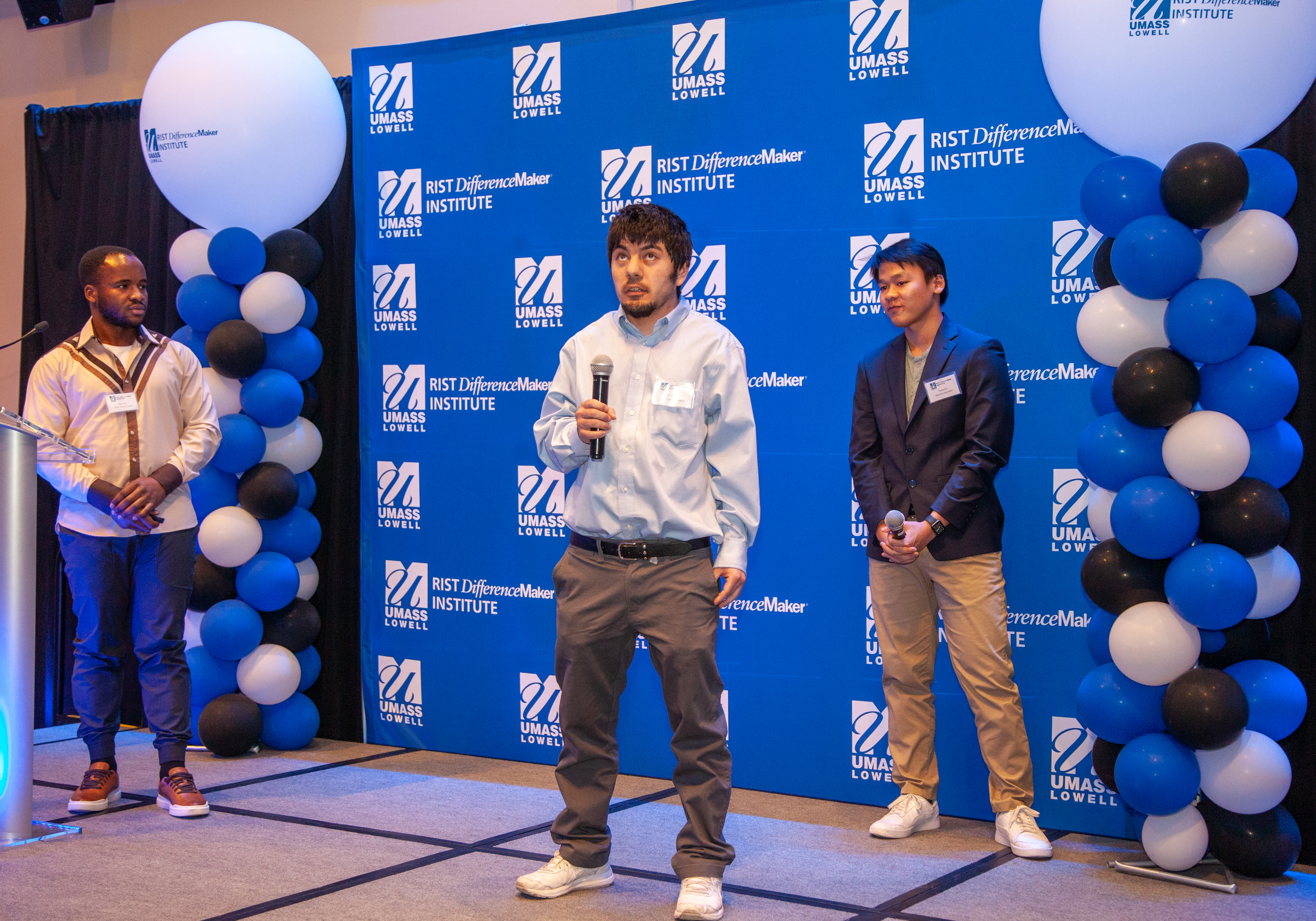 3 male students from the Vital Sensing Patch team speaking and presenting in front of a blue UMass Lowell backdrop and balloons.