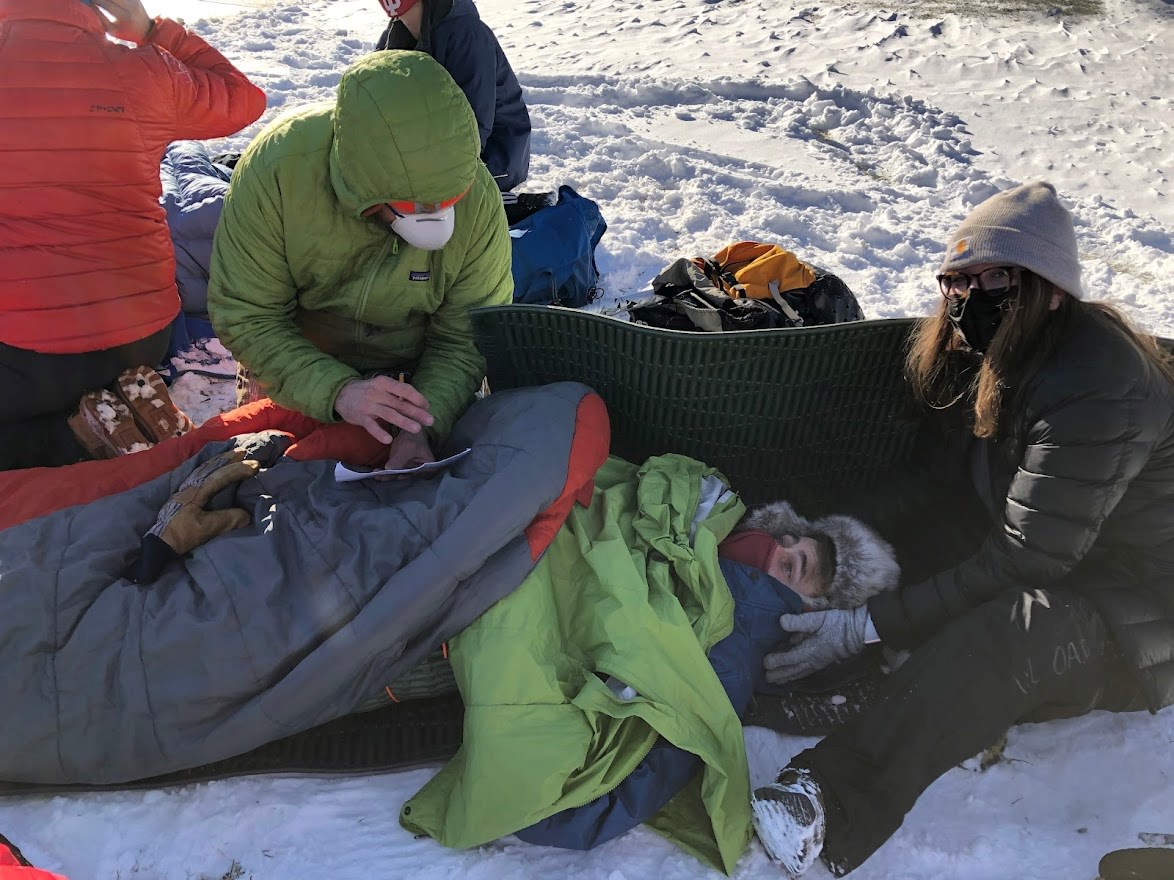 3 people in a practice medical scenario outside, one person lying on a pad with warm layers while two others care for them