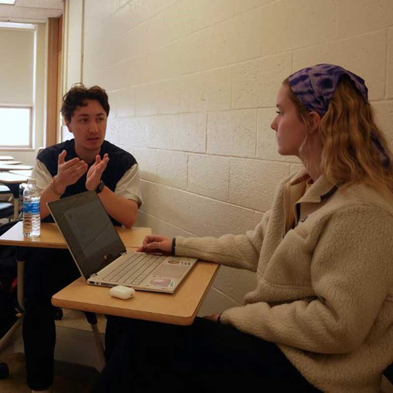 Two students talk while seated in a classroom