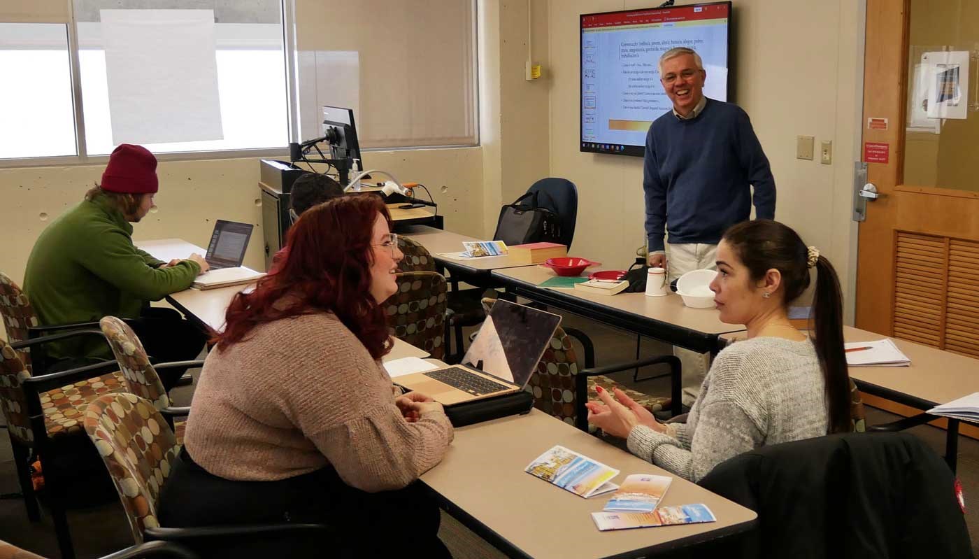 A professor smiles and watches as two students converse in a World Languages and Cultures classroom at UMass Lowell.