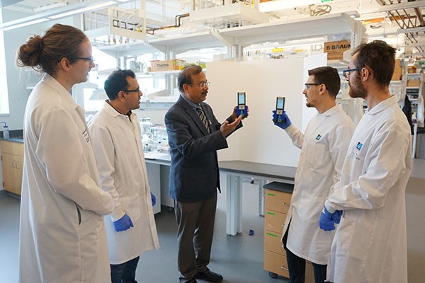 Prof. Pradeep Kurup, center, goes over the functions of the Electronic Tongue surrounded by other researchers in white lab coats.