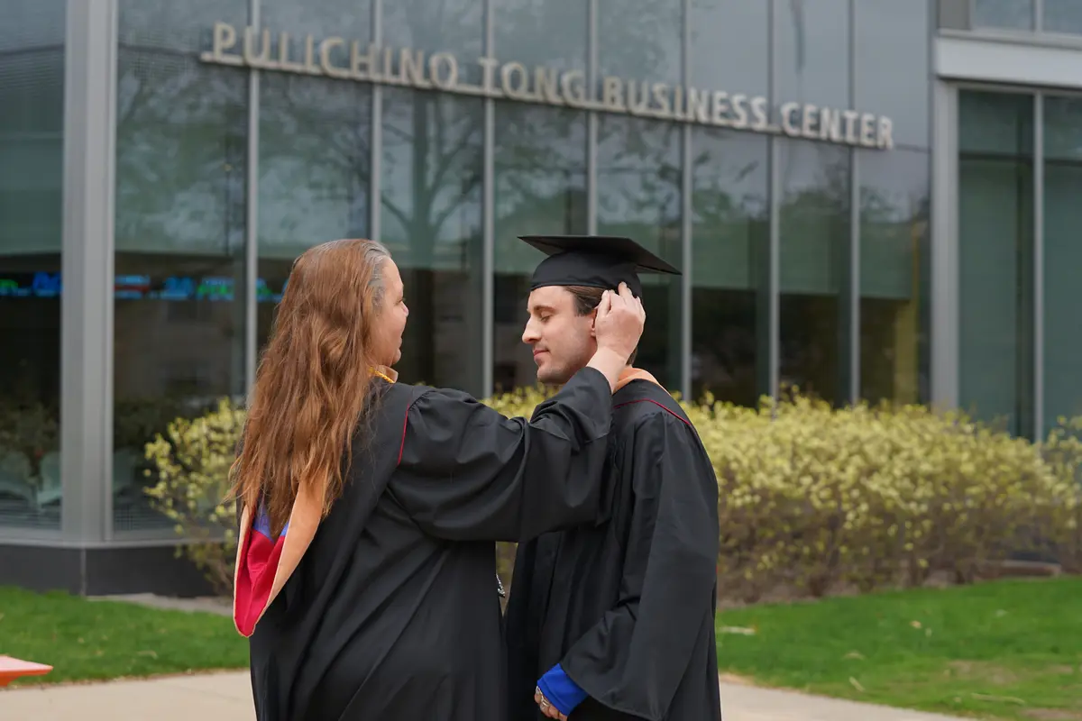 A woman in a college graduation gown helps her son with his regalia while outside a glass building.