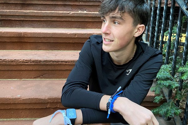 A young man with dark hair sits on a stoop with his arms crossed wearing athletic bracelets