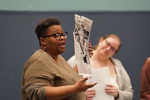 A woman with short hair and glasses holds up a piece of a paper while a woman looks on
