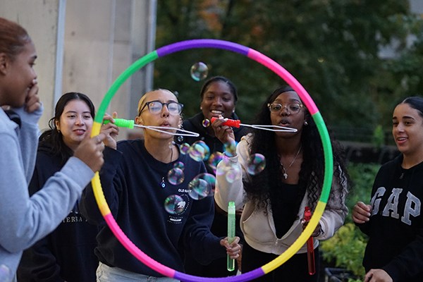 Two young women blow bubbles through a colorful hoop that another woman is holding