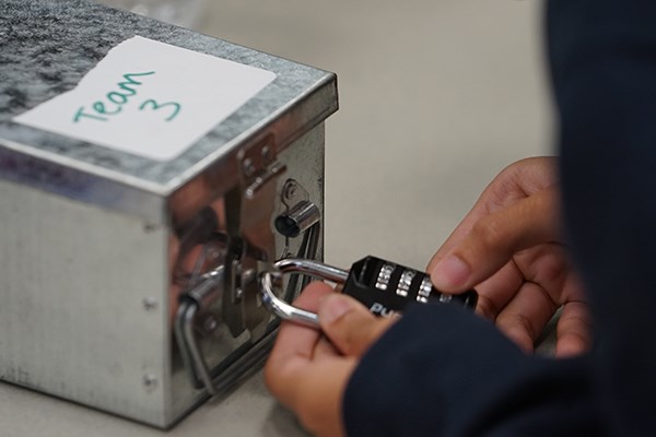 A person works on a combination lock that is attached to a metal box