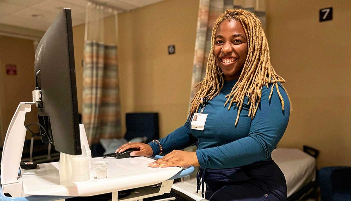 UMass Lowell nursing student Yonnie Collins stands at a computer desk in a hospital room.