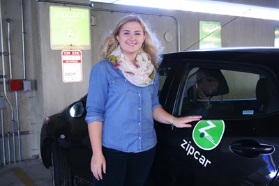 Person next to a black car with the green Zipcar logo parked in a space with signs denoting that the space is reserved solely for Zipcars.