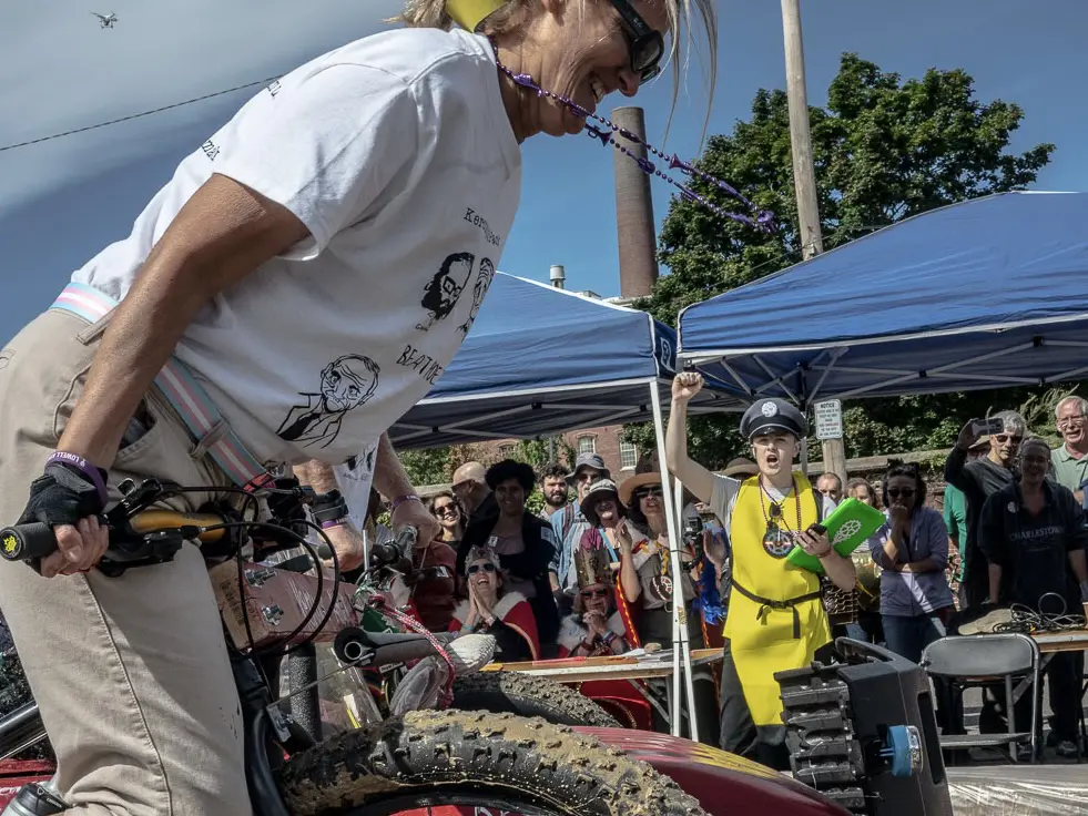 A man in a banana costume and police hat raises his right arm while cheering a woman pedaling through a mud pit.
