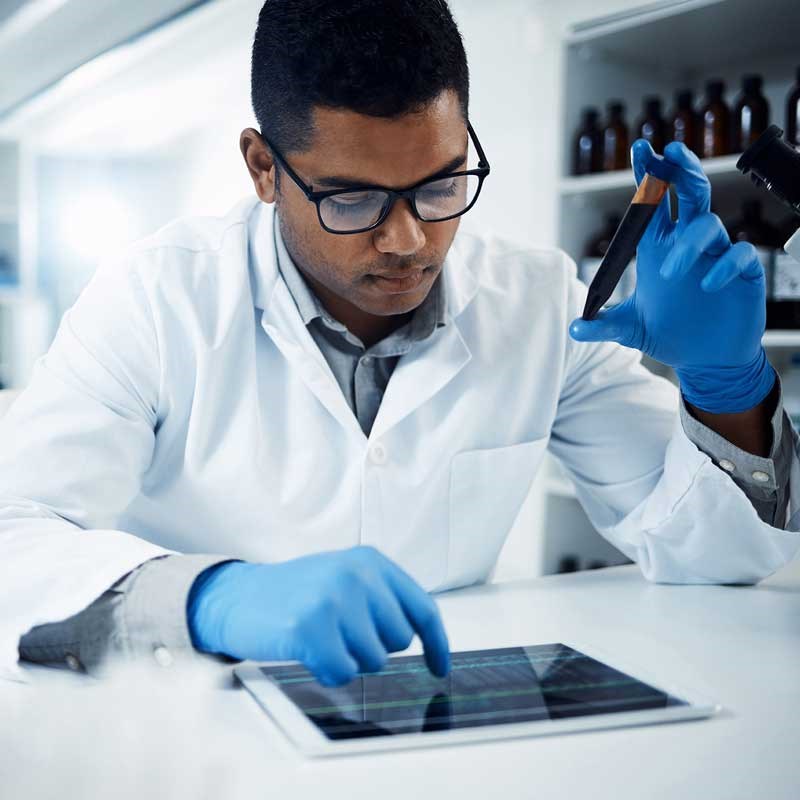 Person wearing a lab coat holds a test tube while looking at a computer tablet