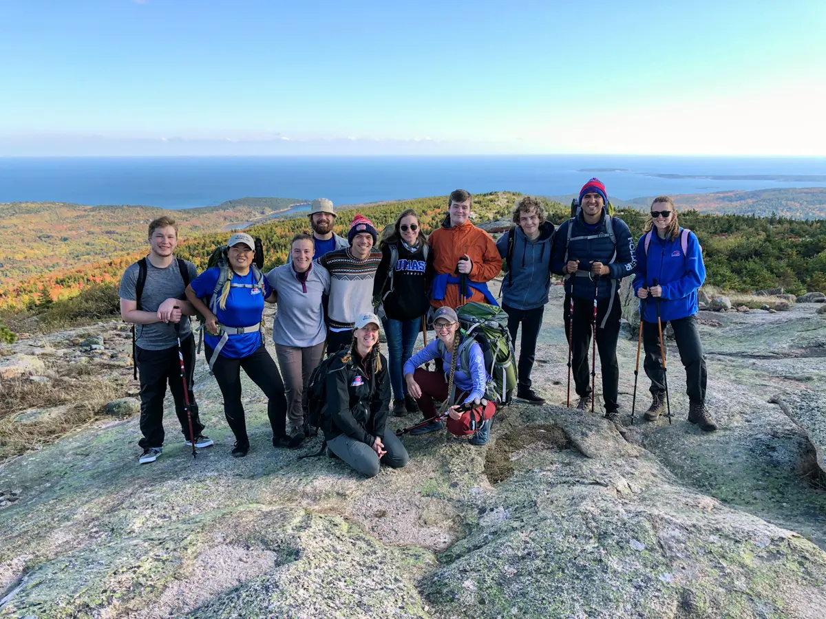 a group of twelve people stand smiling on a rocky outcrop with autumnal trees behind them and the ocean beyond