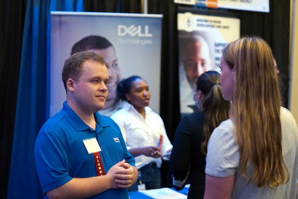 A young man in a blue polo shirt talks to a student at a career fair table