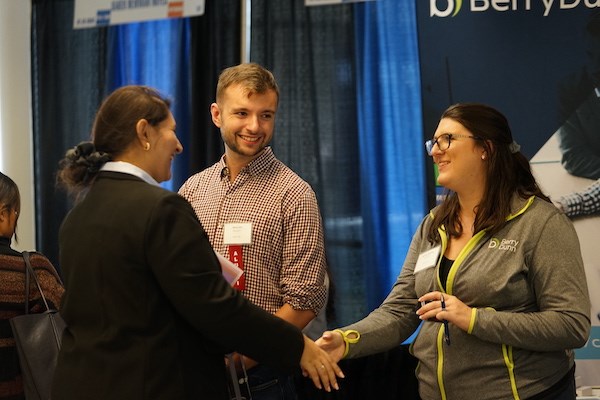 Two women shake hands while a man looks on at a career fair booth