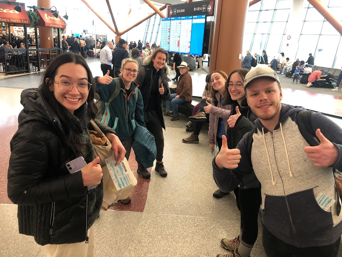 A group of six students stands in an airport terminal holding boarding passes