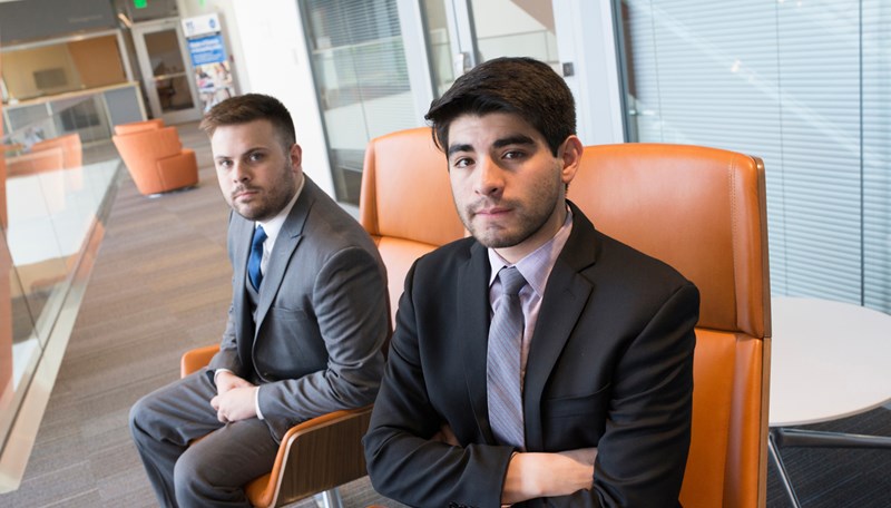 UMass Lowell students and Model U.N. members Alejandro Lopez and Ryan Dekeon, pictured in suits sitting in the Pulichino Tong Building in orange chairs looking at the camera