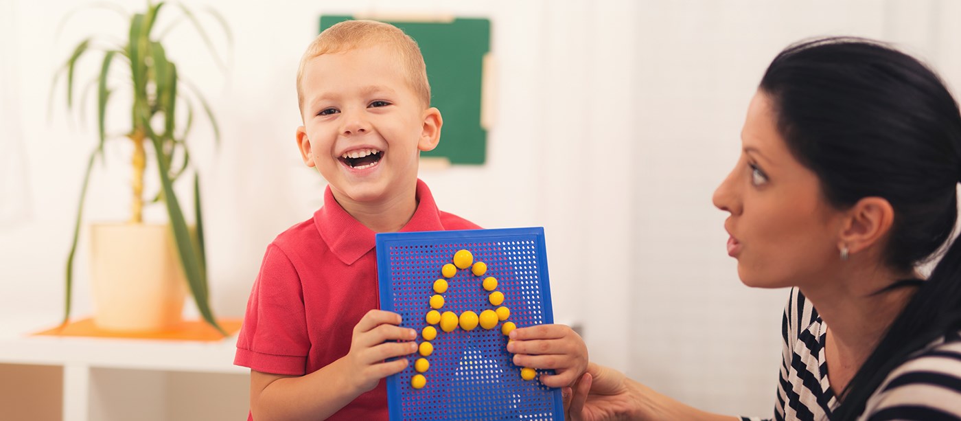 Little boy smiling and holding up letter "A" with female speech therapist. 