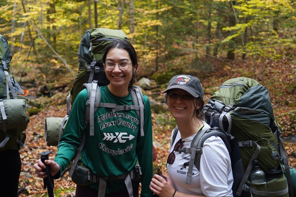Two women smile wearing backpacking in the woods