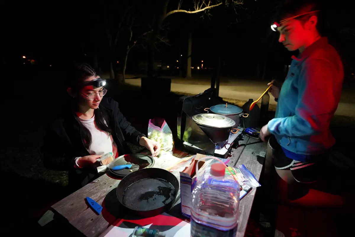 Two people cook on camping stoves in the dark using their headlamps to illuminate the pots and pans.