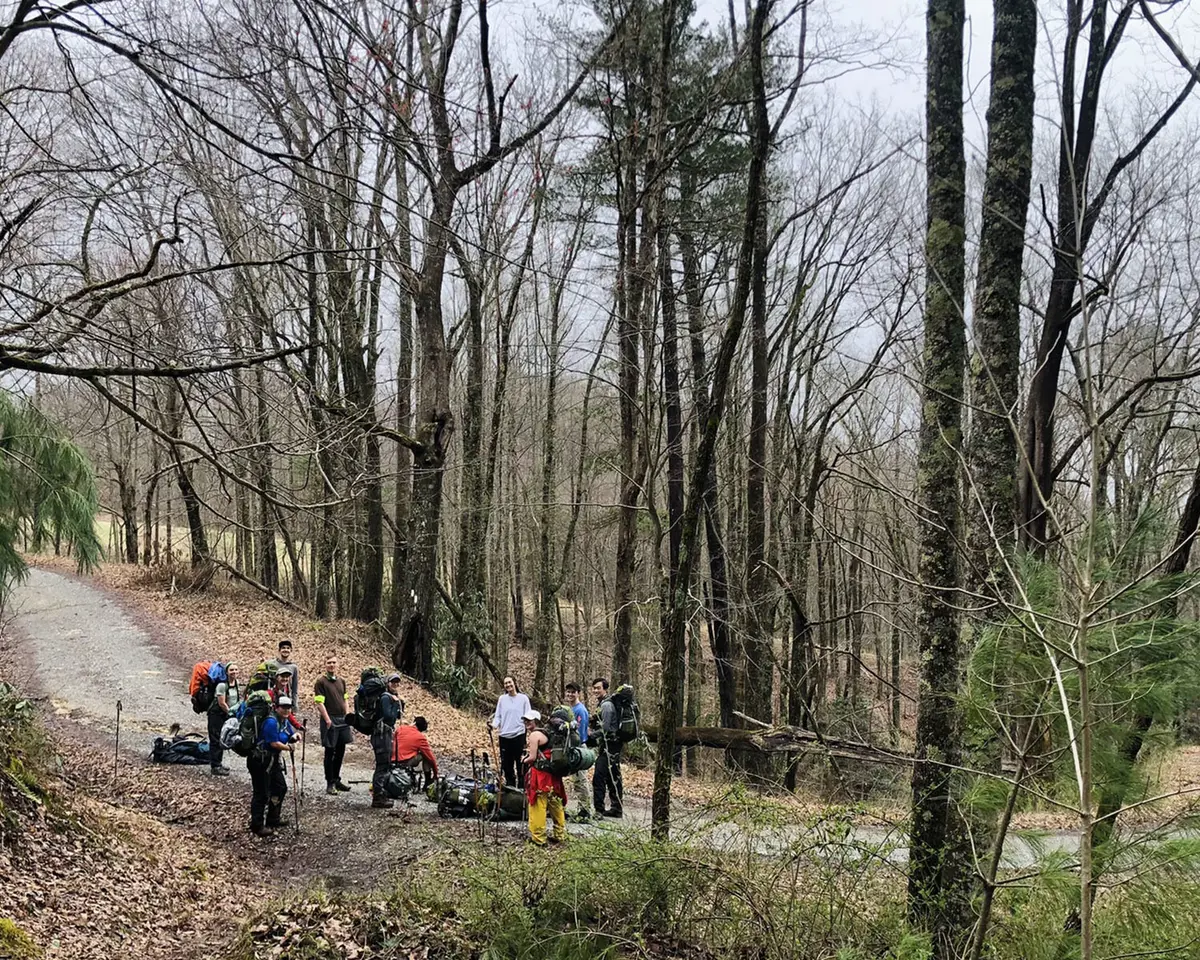 A backpacking group takes a break on the trail amid deciduous trees that haven't yet leafed out. 
