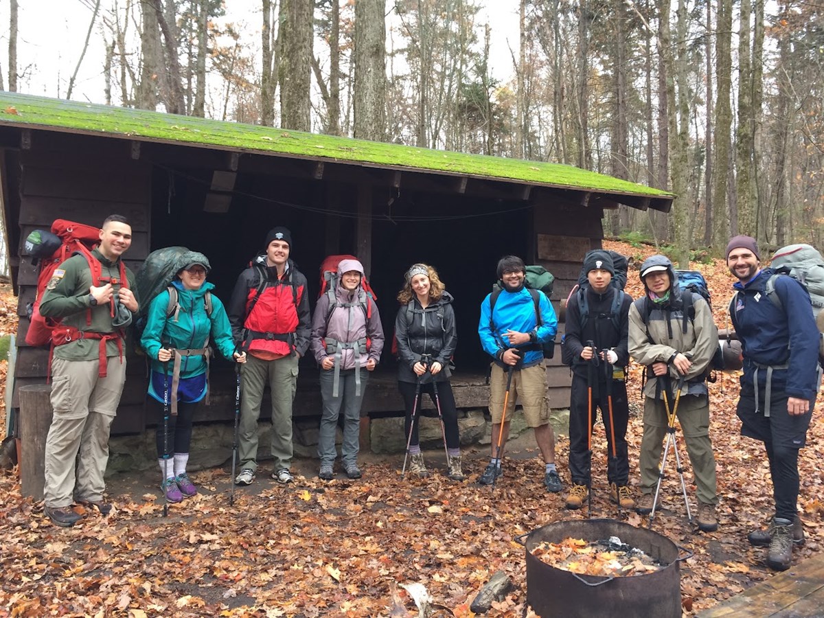 a group of nine stands with backpacking on in front of a shelter, ready to hike